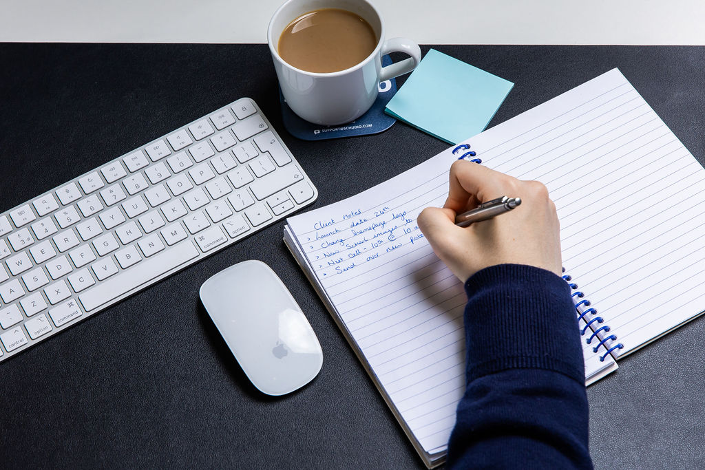 Handwritten planning notes in a notebook beside a keyboard, mouse and cup of tea