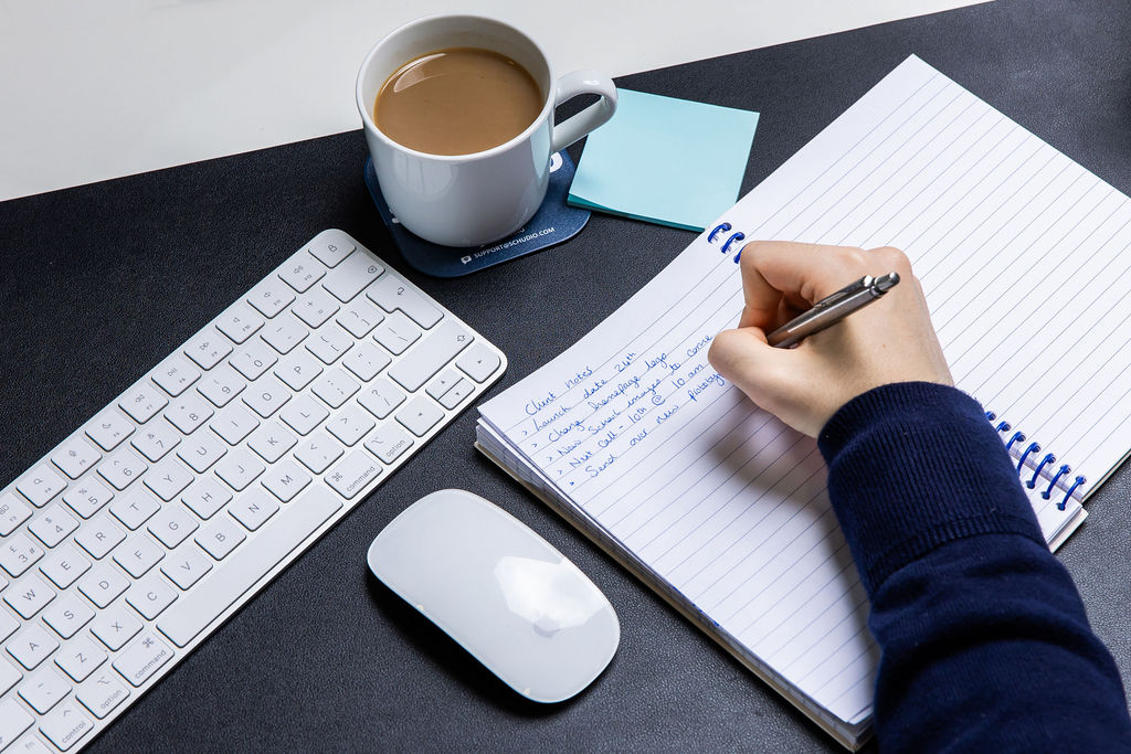 A hand writing a list of school website changes in a notebook beside a keyboard and a Schudio coaster