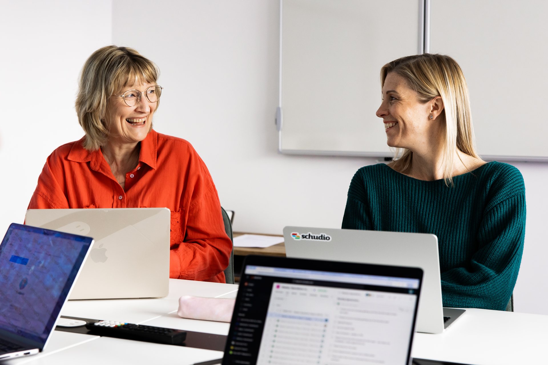 Two members of the Schudio team smiling and talking at a meeting table with laptops open