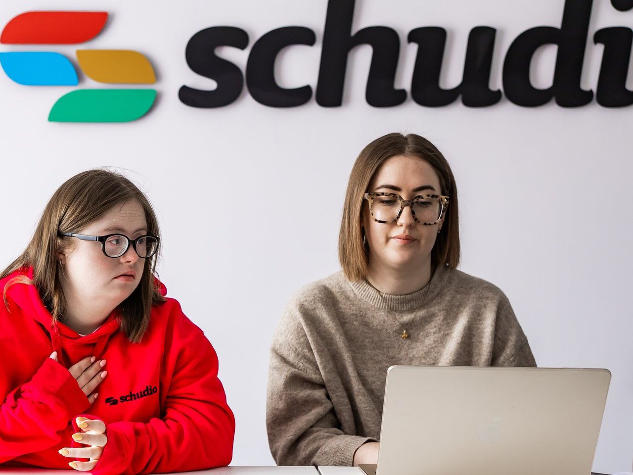 Two Schudio team members at a table in front of the Schudio logo wall, one working on a laptop