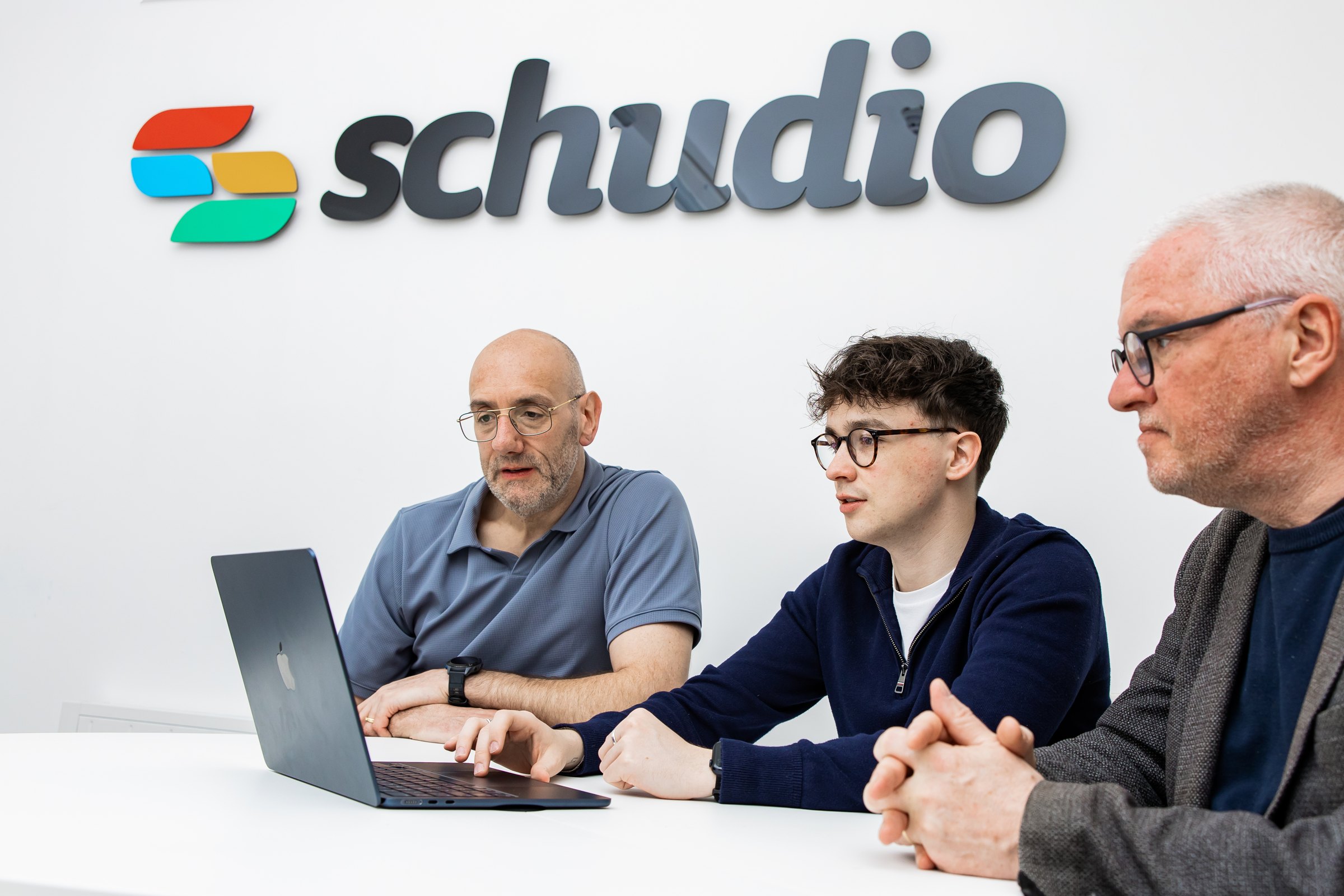 Three members of the Schudio team gathered round a laptop beneath the Schudio logo, talking through the CMS