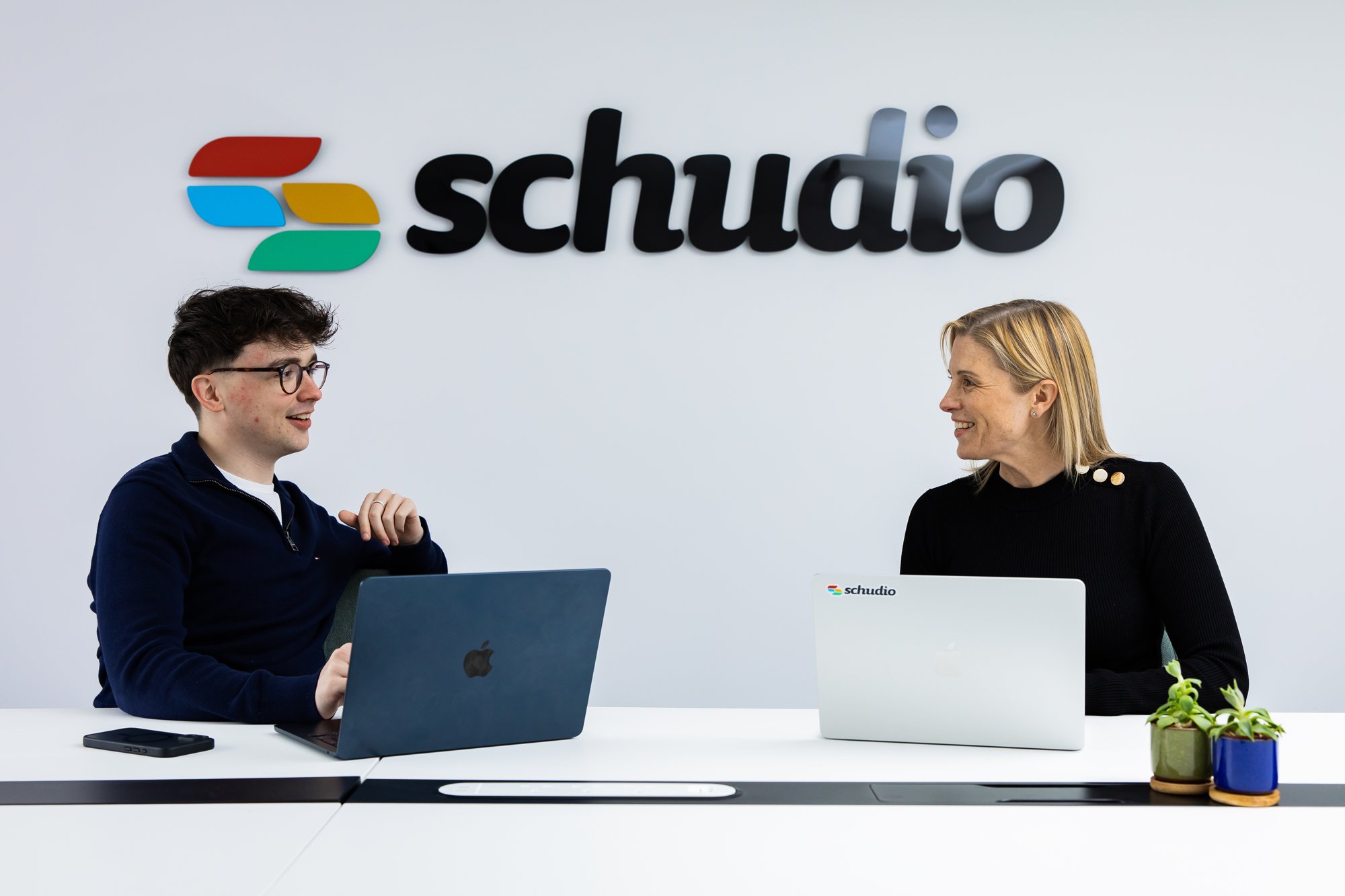 Two Schudio team members talking at laptops in front of the Schudio logo wall